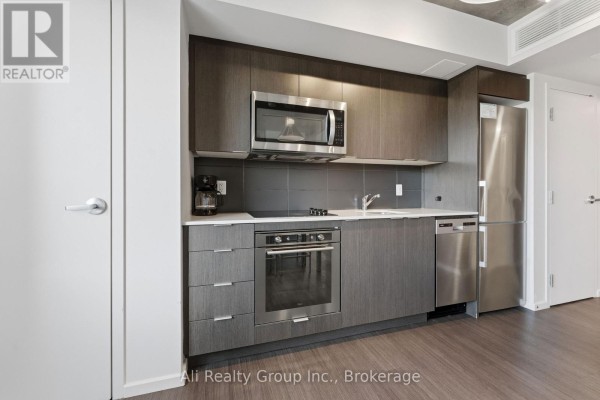 A modern apartment kitchen with dark wood cabinets, stainless steel appliances, and a gray tiled backsplash. - 603 - 105 Champagne Avenue S, Ottawa