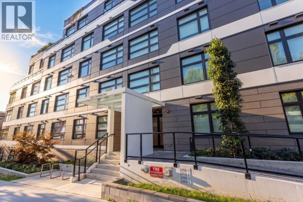 Exterior of a modern apartment building with a covered entrance and landscaping. - 603 3264 Vanness Street, Vancouver