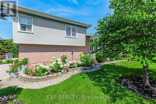 Side view of the house exterior showing the brick base, siding, landscaping with flowers, and a large tree. - 6030 Doane Road, East Gwillimbury
