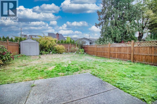 Backyard view with a concrete patio, grassy lawn, wooden fence, and surrounding trees. - 6031 Rumble, Burnaby