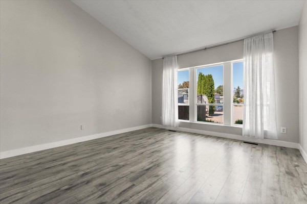 Empty living room with light grey walls, grey wood-look flooring, and large windows with white curtains. - 6033 Glenroy Drive, Sardis South, Chilliwack