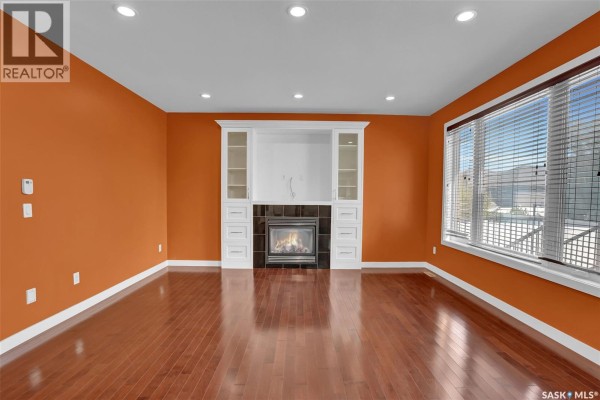 Living room with orange walls, hardwood floors, gas fireplace with built-in shelving, and large window. - 6033 Little Pine Loop, Regina