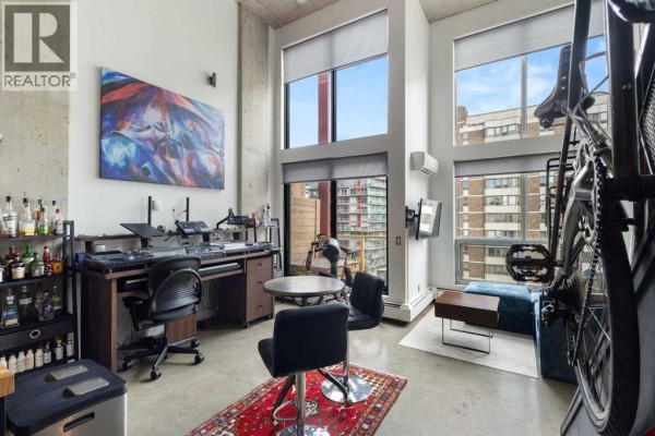 Living space with a large desk setup, exercise bike, seating area, and floor-to-ceiling windows. - 605, 535 8 Avenue Se, Calgary