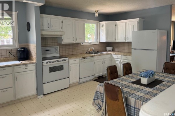 Kitchen with white cabinets, appliances, sink, and a dining table with a checkered tablecloth. - 605 5th Avenue W, Assiniboia