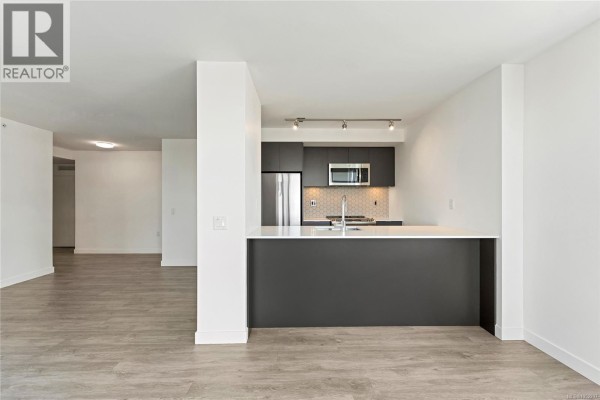 Living room and kitchen area with large windows showing urban buildings and grey flooring. - 608 845 Johnson St, Victoria