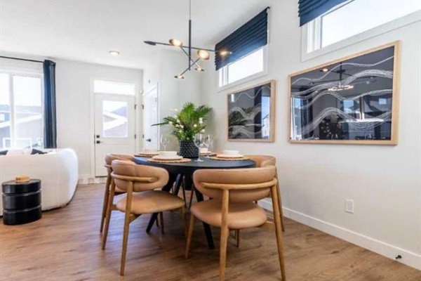 Dining area with a round black table, four tan chairs, and modern wall art. A large window and door are visible. - 609 Lambeau Link Se, Airdrie