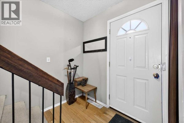 Entranceway with a white door, hardwood floors, and a staircase with a wooden railing. - 61, 4940 39 Avenue Sw, Calgary