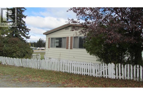 Side exterior of a beige mobile home with a white picket fence and trees. - 61 375 Horse Lake Road, 100 Mile House