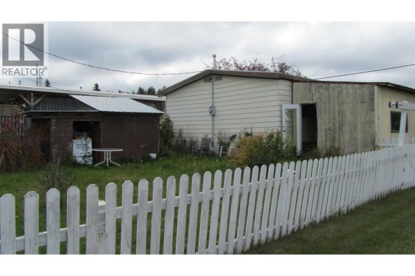 Rear exterior of a mobile home with a detached shed, white picket fence, and lawn. - 61 375 Horse Lake Road, 100 Mile House
