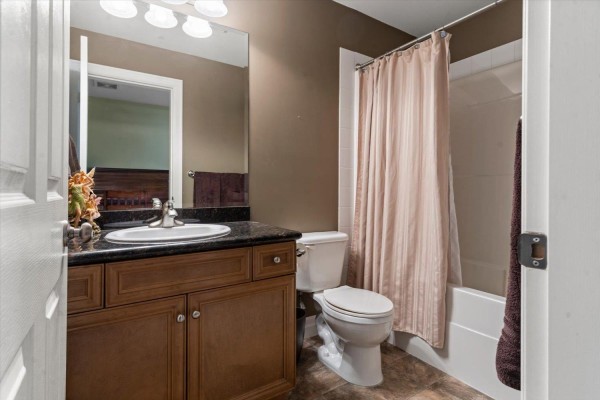 A bathroom with a brown vanity, granite countertop, toilet, and a bathtub with a beige shower curtain. - 61 6449 Blackwood Lane, Sardis South, Chilliwack