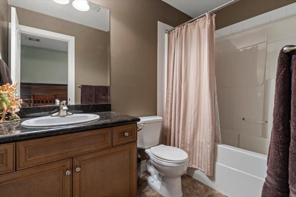 A bathroom with a brown vanity, granite countertop, toilet, and a bathtub with a beige shower curtain. - 61 6449 Blackwood Lane, Sardis South, Chilliwack