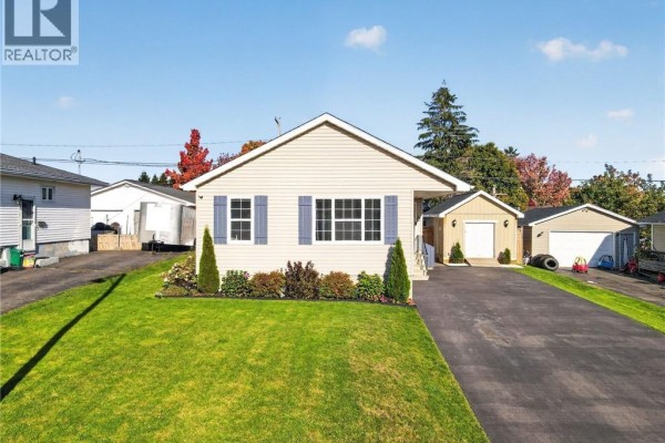Front view of a beige vinyl-sided bungalow with a black roof, blue shutters, and attached garage. - 61 Bolton Street, Moncton