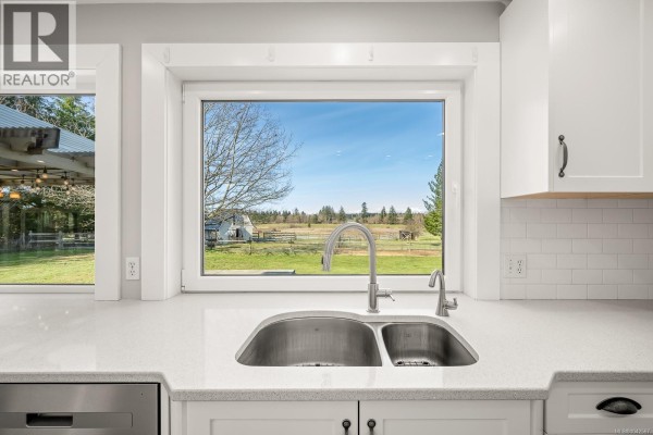 Interior view of a living area with a brick fireplace, large windows, sliding doors, and kitchen counter in background. - 6104 Ledingham Rd, Courtenay
