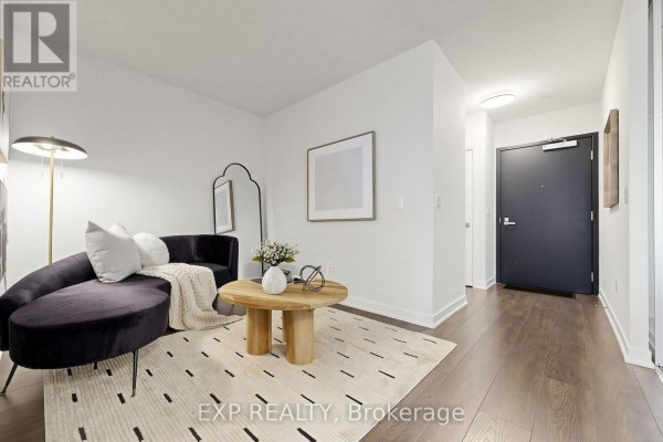 Living room area with dark sofa, coffee table, floor lamp, and entrance hallway. - 611 - 31 Tippett Road, Toronto