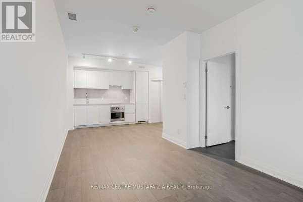 Living area with white walls, grey wood floors, and an open-concept kitchen. - 611 - 4015 The Exchange, Mississauga