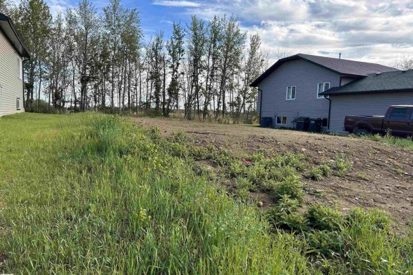 Overgrown grassy lot with a beige house and trees behind the cleared area. - 6111 53a Av, Redwater