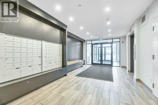Apartment building lobby with white mailboxes, dark grey accent wall, seating bench, and glass entrance doors. - 6112, 200 Seton Circle Se, Calgary