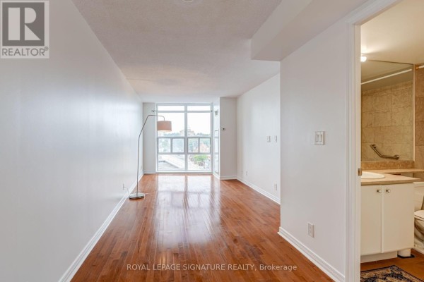 Living room with hardwood floors, large window, and a floor lamp. Bathroom entrance visible. - 612 - 736 Spadina Avenue, Toronto