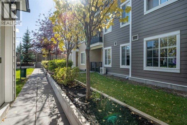 Stone pathway beside a landscaped garden bed with trees and grass, with exterior of townhome building. - 6121, 151 Legacy Main Street Se, Calgary