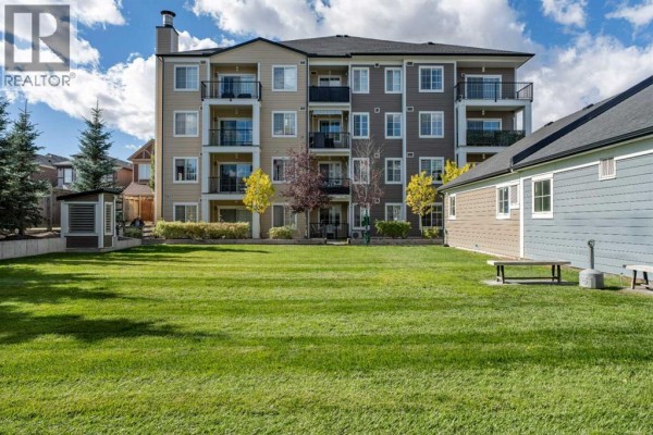 Exterior of a multi-story apartment building with beige and grey siding, balconies, and a large grassy lawn. - 6121, 151 Legacy Main Street Se, Calgary