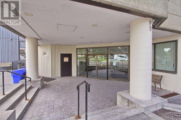 Covered building entrance with beige tiled floor, glass doors, and concrete pillars. - 613 - 5580 Sheppard E Avenue, Toronto