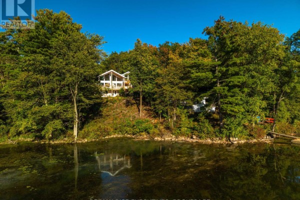 Home with extensive windows nestled amongst mature trees on a wooded hillside overlooking a calm lake. - 62 Apple Jack Lane, Frontenac