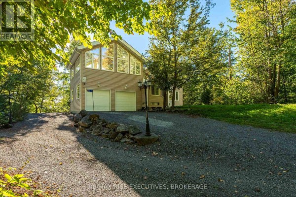 Two-car garage doors visible on a tan home with a gravel driveway and stone landscaping. - 62 Apple Jack Lane, Frontenac