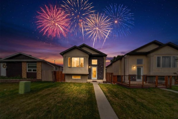 Night exterior view of a modern Winnipeg home with illuminated windows and fireworks in the sky. - 62 La Porte Drive, Winnipeg