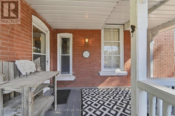 Covered porch with a black and white patterned rug, brick walls, and a rustic wooden bench. - 62 Orange Street S, Central Huron