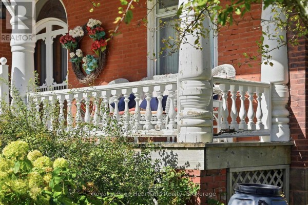 White ornate porch railing with decorative columns and surrounding green foliage on a brick house. - 62 Orange Street S, Central Huron