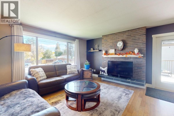 Living room with a brick fireplace, television, large window overlooking the front yard, and leather sofas. - 620 Ziprick Road, Kelowna