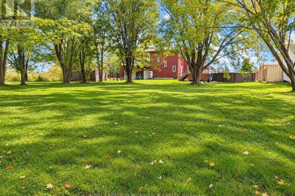 Large, sun-dappled lawn with mature trees, showing a red-sided building and swimming pool in the distance. - 6208 165e Avenue, South Glengarry