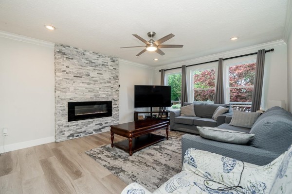 Living room with grey sectional sofa, fireplace with stacked stone surround, wood coffee table, and large windows. - 6209 172 Street, Surrey