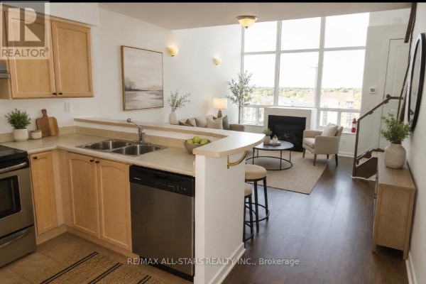 Kitchen counter with double sink, dishwasher, and view of living area with fireplace and large windows. - 621 - 250 Manitoba Street, Toronto