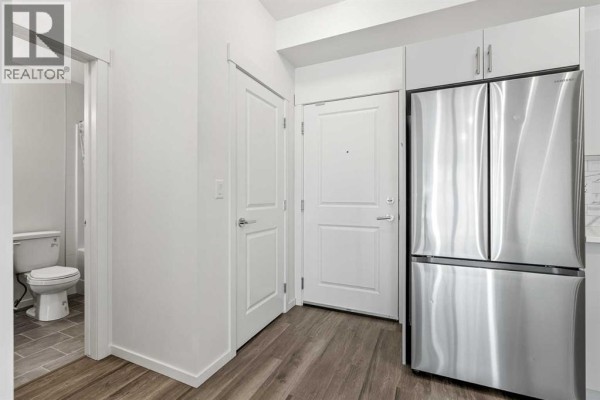 Interior view of an apartment hallway with a stainless steel refrigerator and a bathroom door. - 6215, 15 Sage Meadows Landing Nw, Calgary