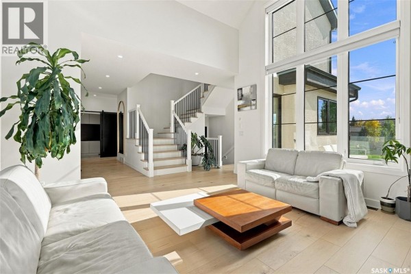 Open concept living area with white sofas, staircase, and large windows overlooking the yard. - 6221 Koep Avenue, Regina