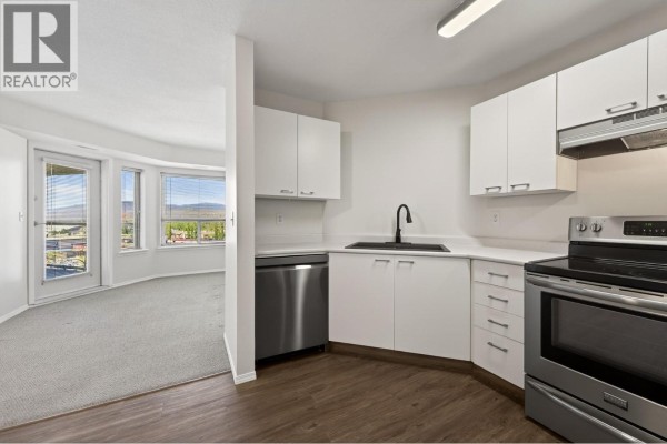 Kitchen area with white cabinets, stainless steel appliances, and a view into a living room with windows. - 629 Lansdowne Street Unit# 605, Kamloops