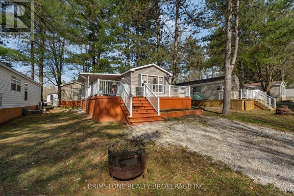 A grey mobile home with a deck and stairs, nestled among mature trees. - 63 - 1082 Shamrock Marina Road, Gravenhurst