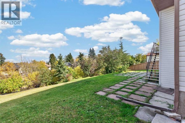 Looking out over the backyard with a large green lawn, paved patio, and distant trees. - 63 Beaconsfield Crescent Nw, Calgary