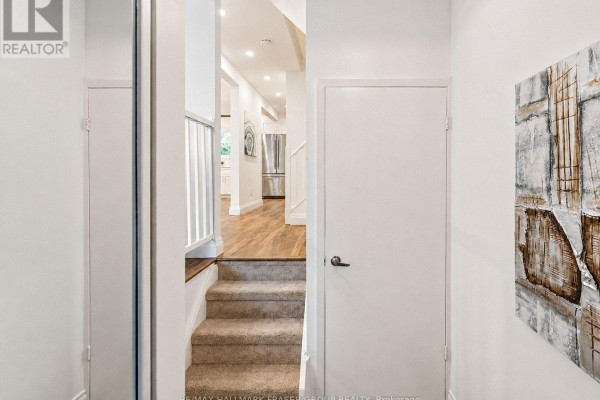 Interior view of a hallway with stairs leading up, beige carpet, white doors, and a glimpse of a kitchen. - 63 Willows Lane, Ajax