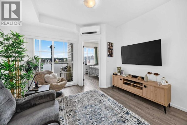 Living room featuring a grey sofa, wooden media console, television, and a sliding glass door to a balcony. - 6305, 151 Legacy Main Street Se, Calgary