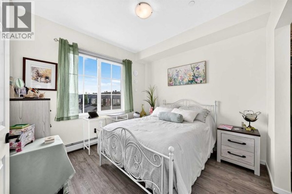 Bedroom with a white metal bed frame, a desk by the window, and a view of the neighbourhood. - 6305, 151 Legacy Main Street Se, Calgary