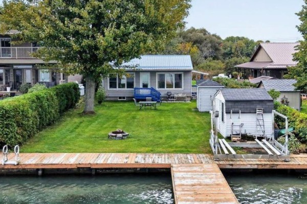 Rear view of a grey cottage with a blue deck, patio furniture, and a wooden dock with boat lift. - 6325 78th Avenue, South Glengarry