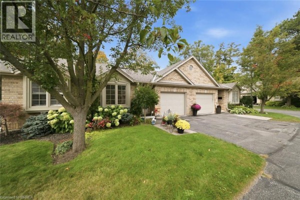 Close-up exterior view of a brick townhouse unit with green lawn and mature trees. - 633 Park Road N Unit# 8, Brantford