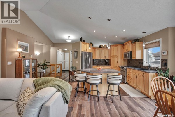 Open concept living area with a kitchen island, light wood cabinets, stainless steel appliances, and living room furniture. - 634 Allwood Manor, Saskatoon