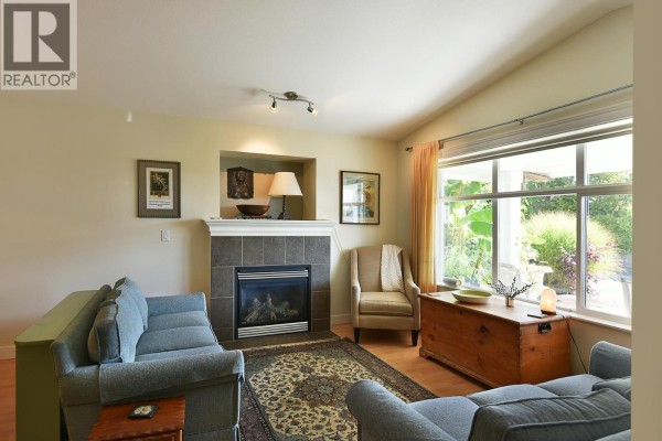 Living room with grey sofas, a tiled fireplace with gas insert, armchair, and a large window. - 6340 Williams Place, Sechelt