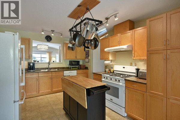 Kitchen with light wood cabinets, white gas stove, dark island on wheels, and a hanging pot rack. - 6340 Williams Place, Sechelt