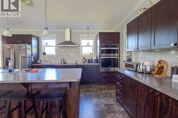 Kitchen view showing dark wood cabinets, white tile backsplash, stainless steel appliances including double ovens, and a granite countertop. - 6380 Dawsons Lane, Smithers