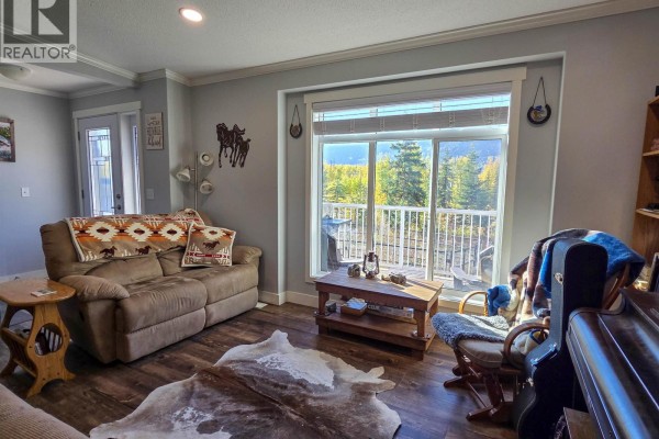 Living room with a brown sofa, rocking chair, coffee table, large window with mountain view, and a cowhide rug. - 6380 Dawsons Lane, Smithers