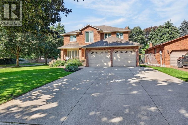 Two-story red brick house with a large concrete driveway and mature trees. - 64 Driftwood Court, Grimsby